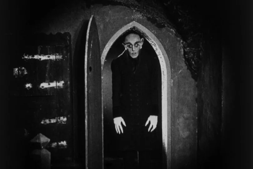 A pale figure stands inside a pointed stone doorway, surrounded by deep shadow.