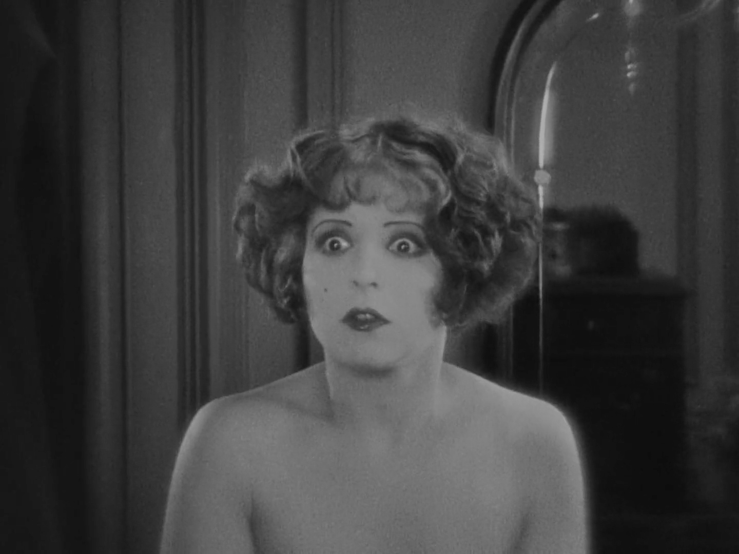 A woman looks forward, her face and shoulders bright against a dark wall and mirror.